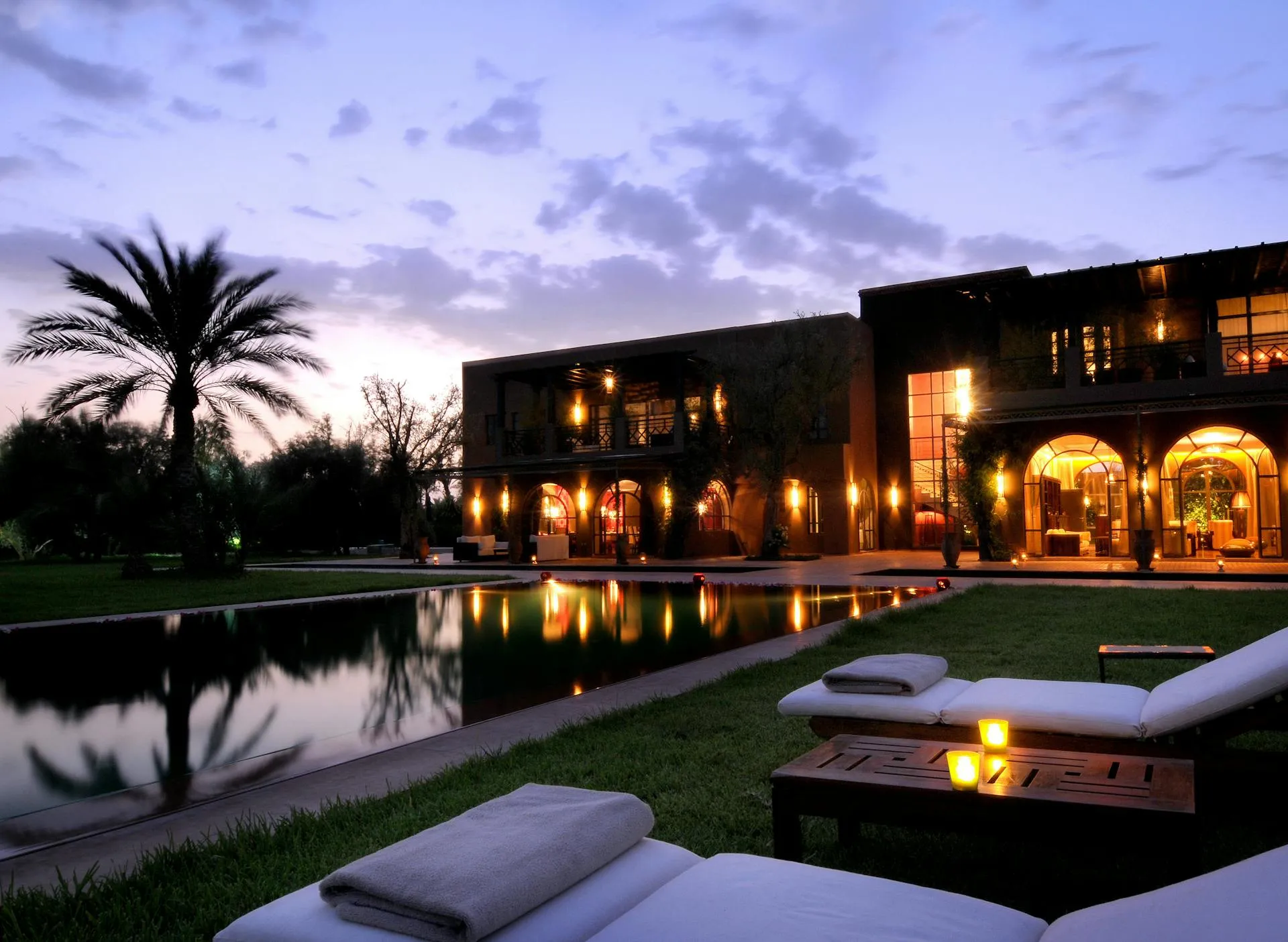A luxury villa glowing at dusk, its lit arches and pool reflecting against a blue-hour sky