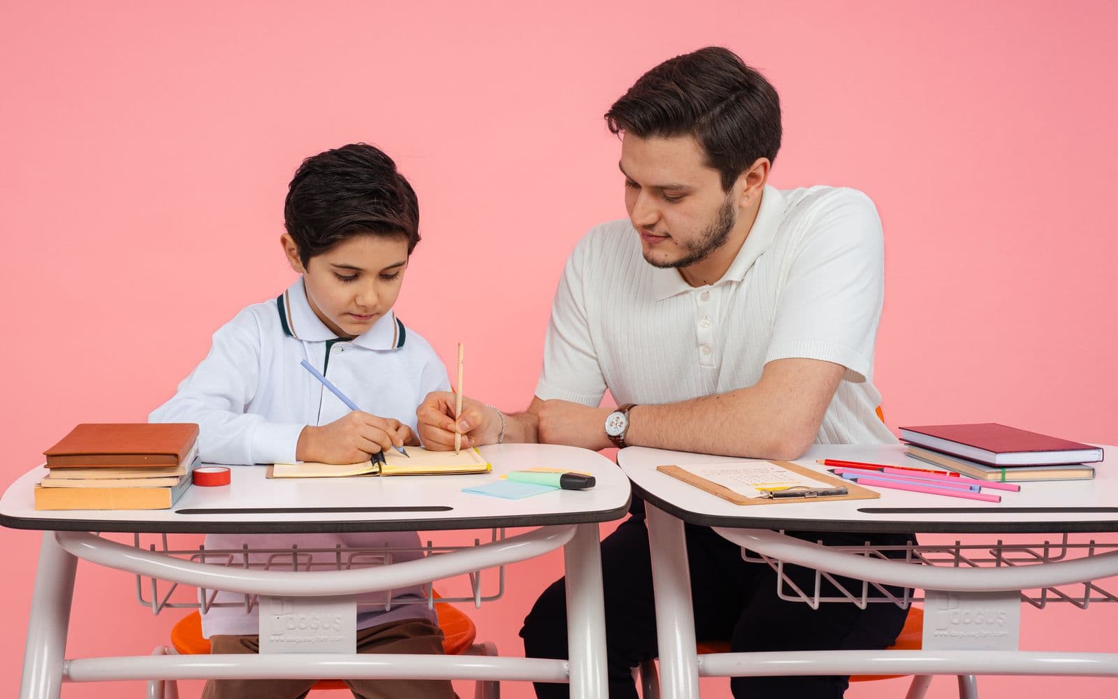 A young boy writing in a notebook at a school desk while an adult sits beside him and helps