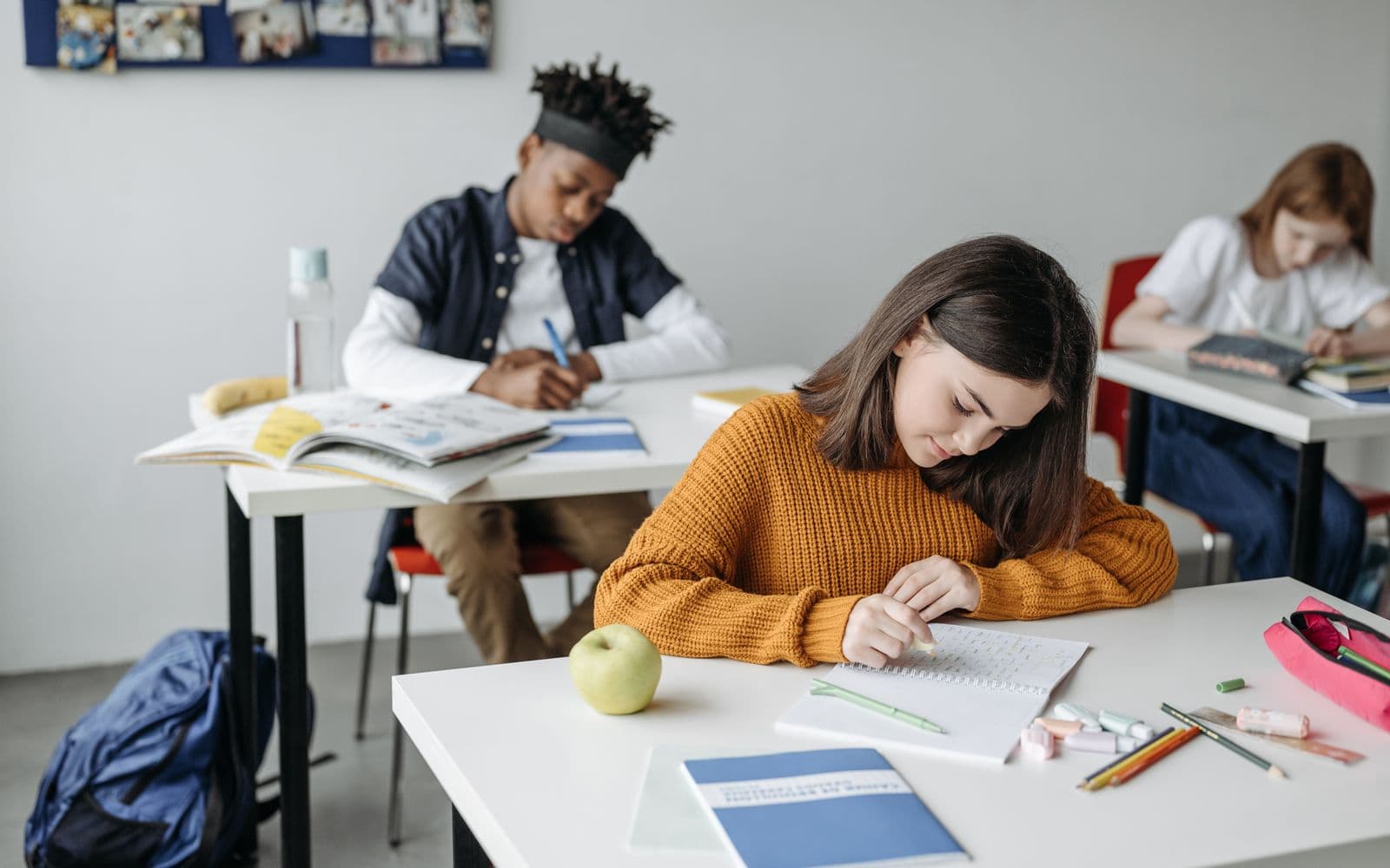 A secondary school student highlighting handwritten notes in a spiral notepad at her classroom desk, with classmates working behind her