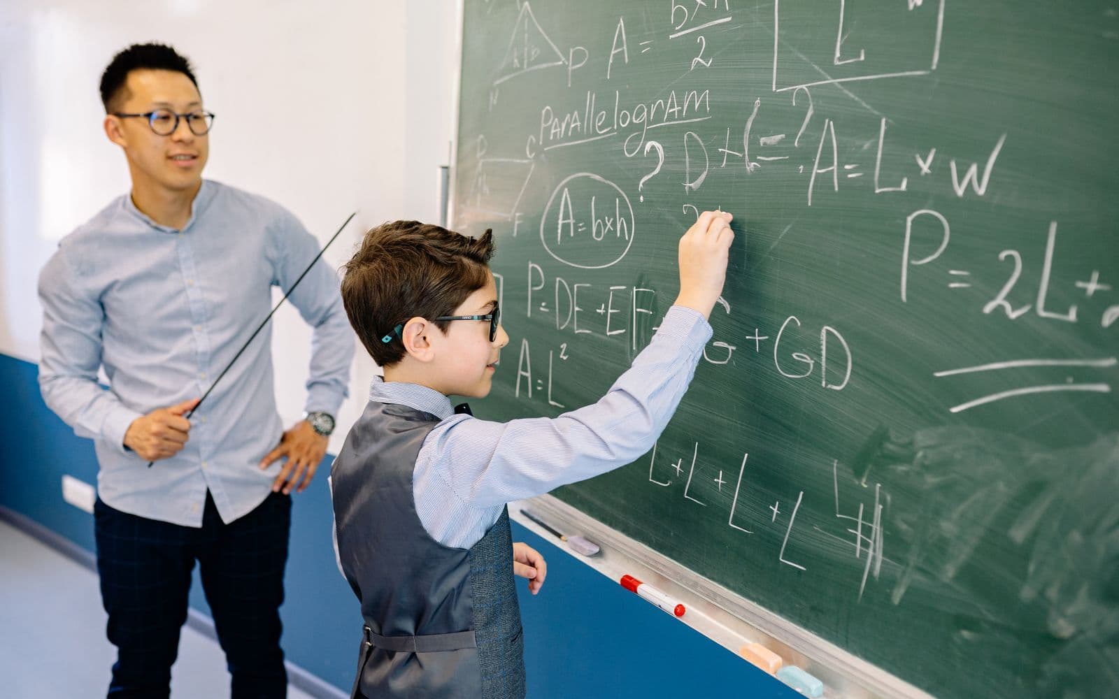 A teacher guides a student solving math equations on a classroom chalkboard.