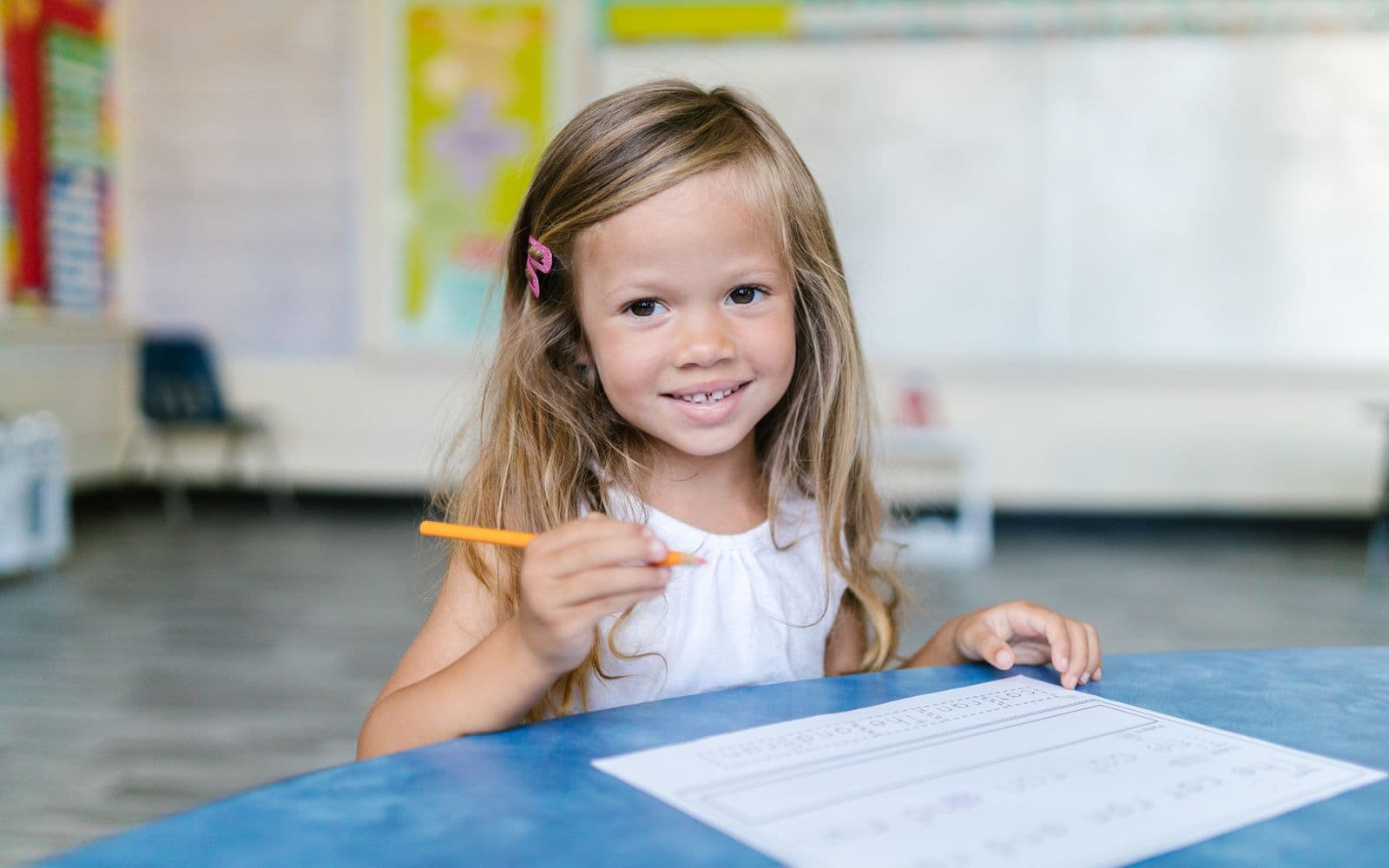 A young girl smiles while working on schoolwork in a bright classroom.
