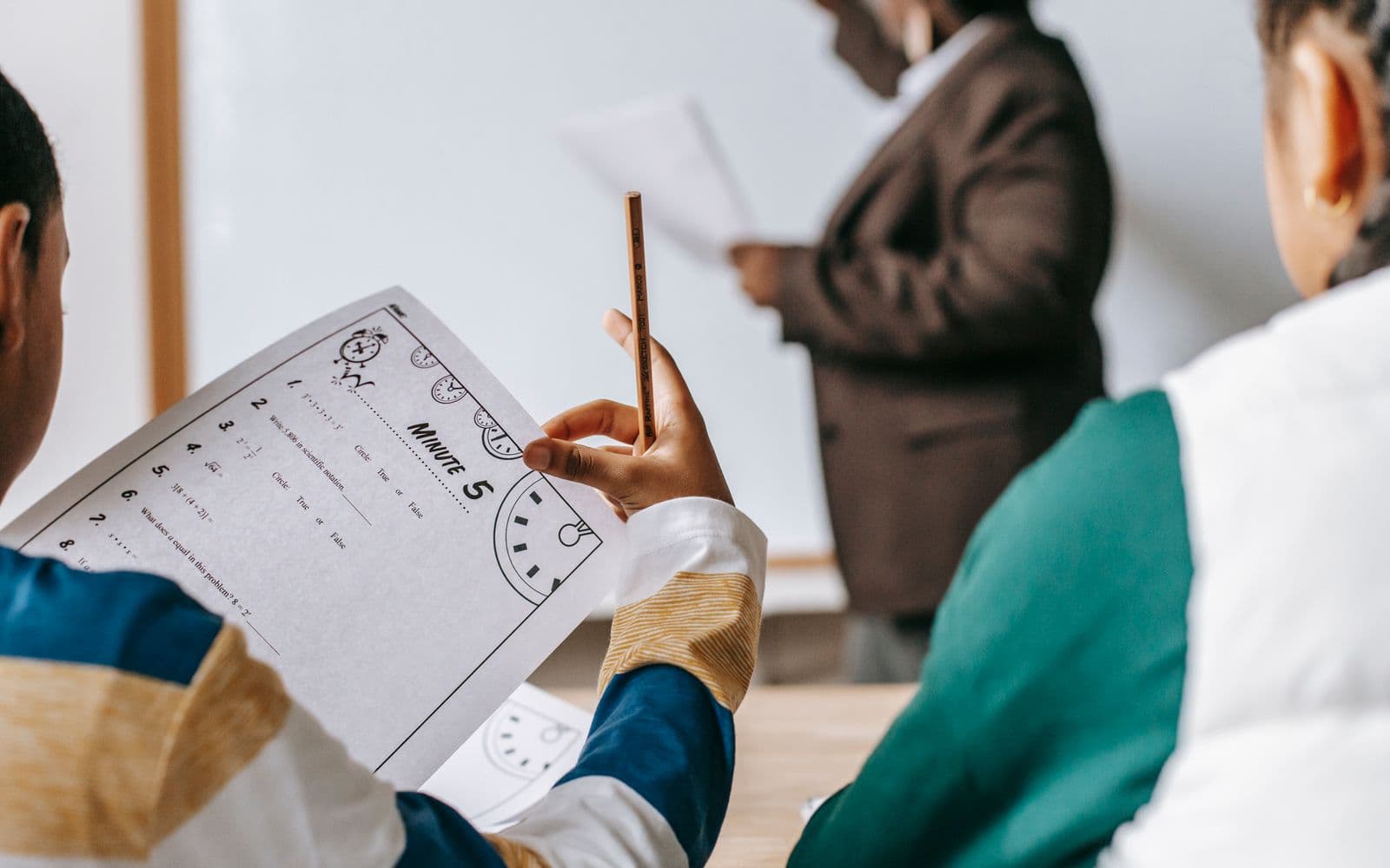 Multiethnic elementary classmates sitting at desk with copybooks and listening to black female teacher writing on whiteboard