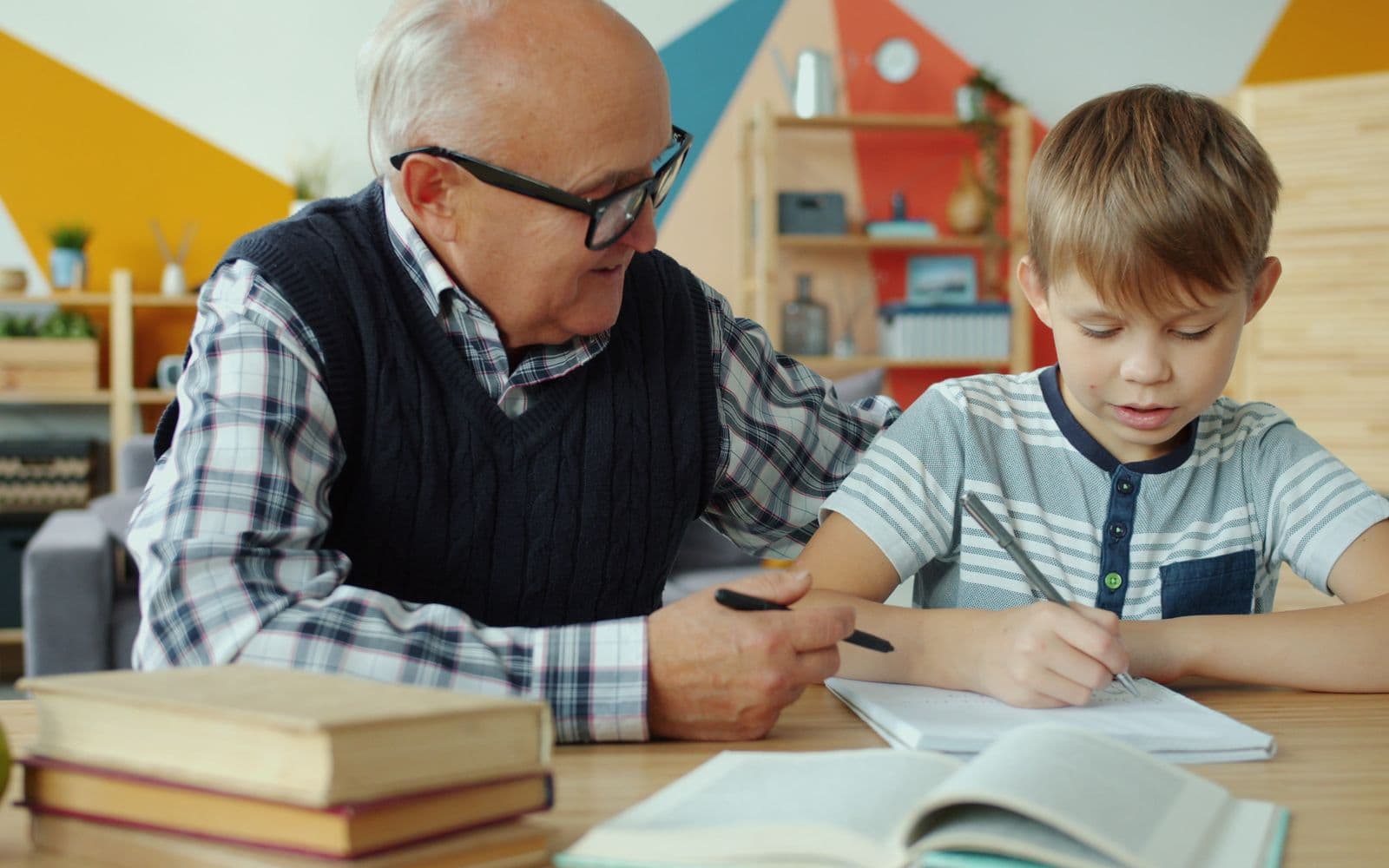 An older man pointing at an exercise book while a boy writes at a table stacked with textbooks