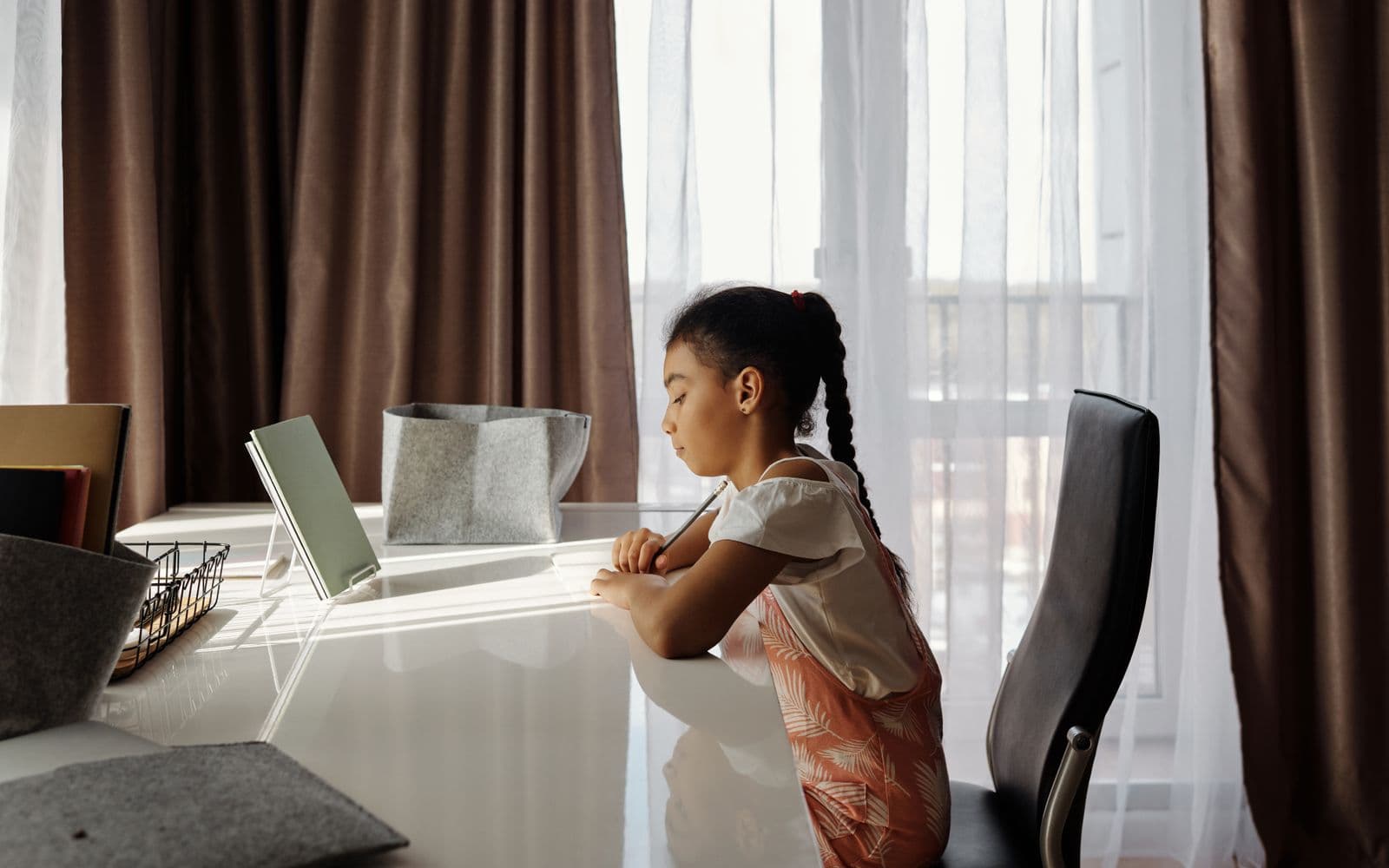 A girl working with a pencil at a desk by a sunlit window, a book propped open on a stand