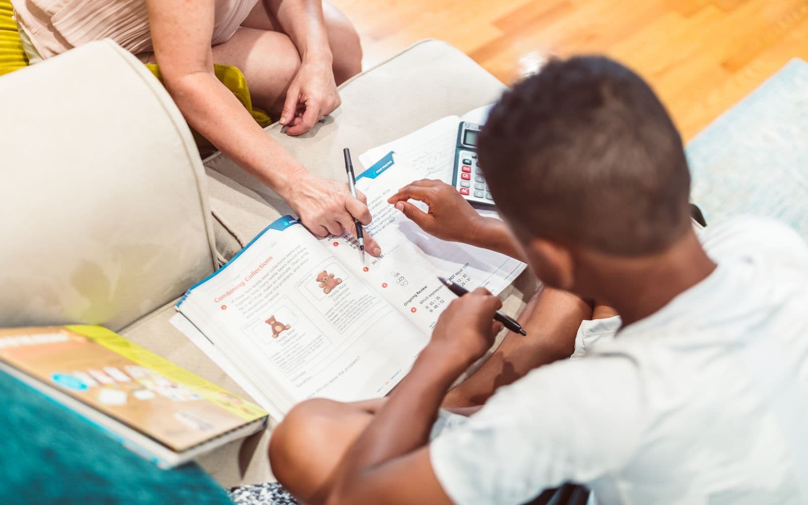 An adult and a child working through a page of a primary maths practice workbook together, with a calculator on the table beside them