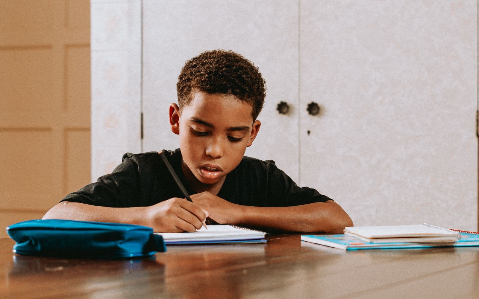 A boy of about ten writing in an exercise book at a table, a pencil case and school books beside him