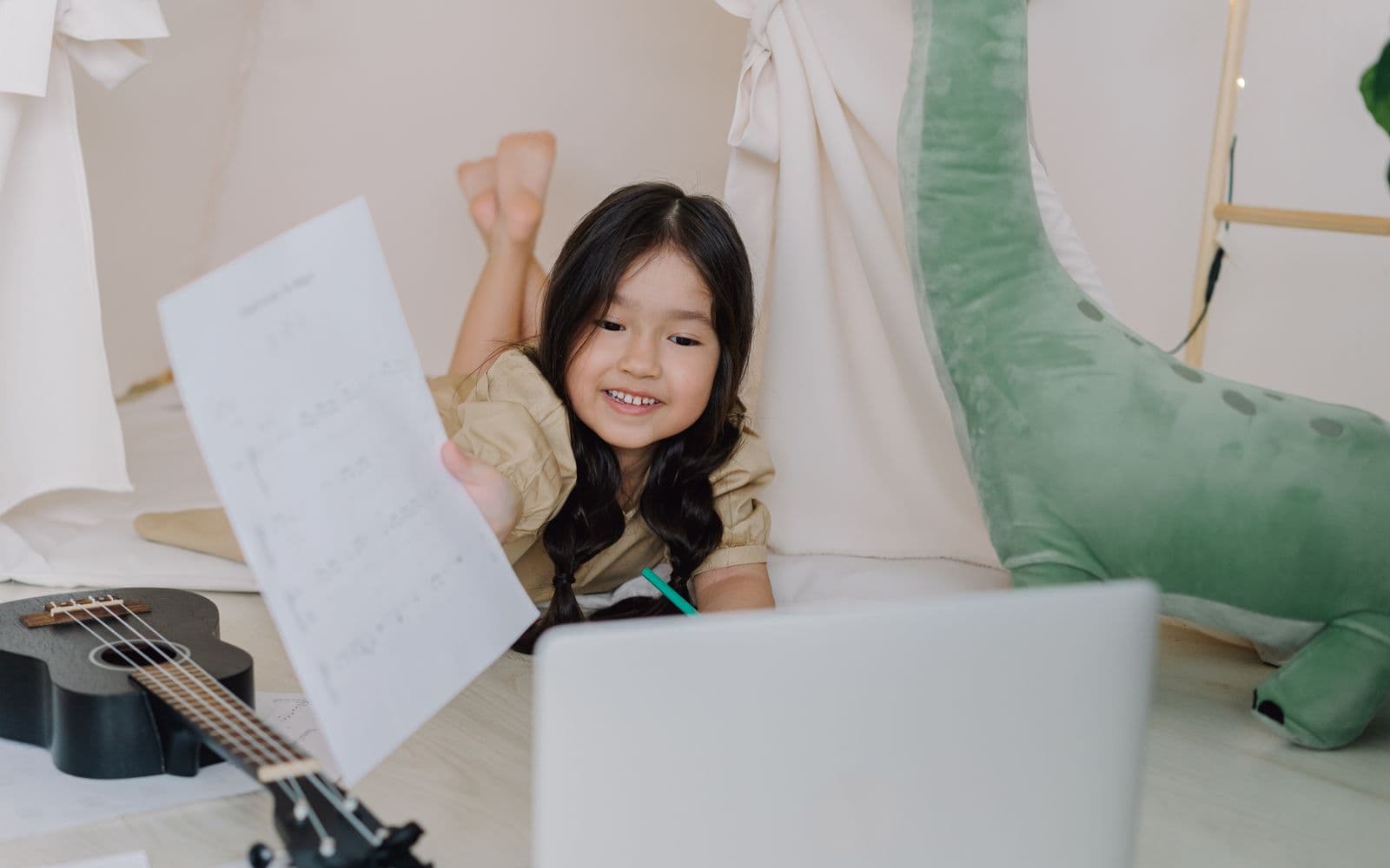 A child at home following an online lesson on a laptop, holding a practice sheet