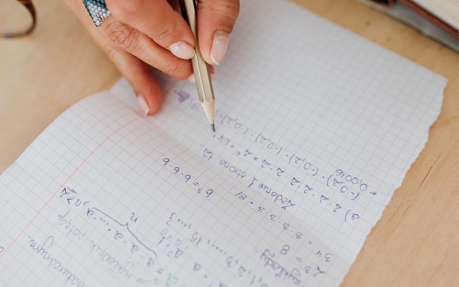 A person writes mathematical equations by hand on grid paper using a pencil, promoting learning.