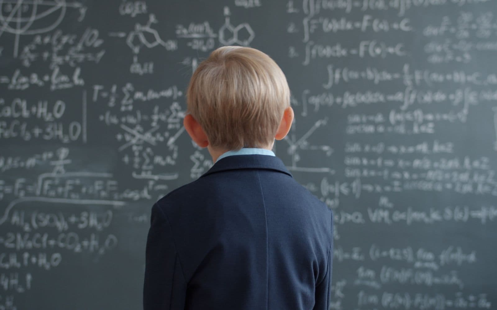 Child in school uniform studying complex math on chalkboard.